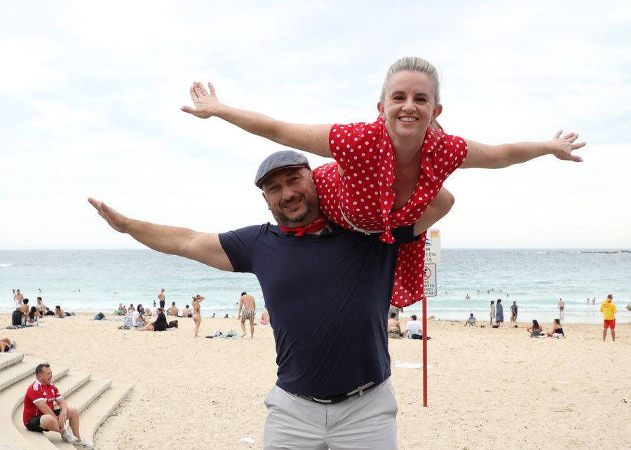 Mel lifted across Marc's shoulders at the beach, both with arms outstretched and grinning.