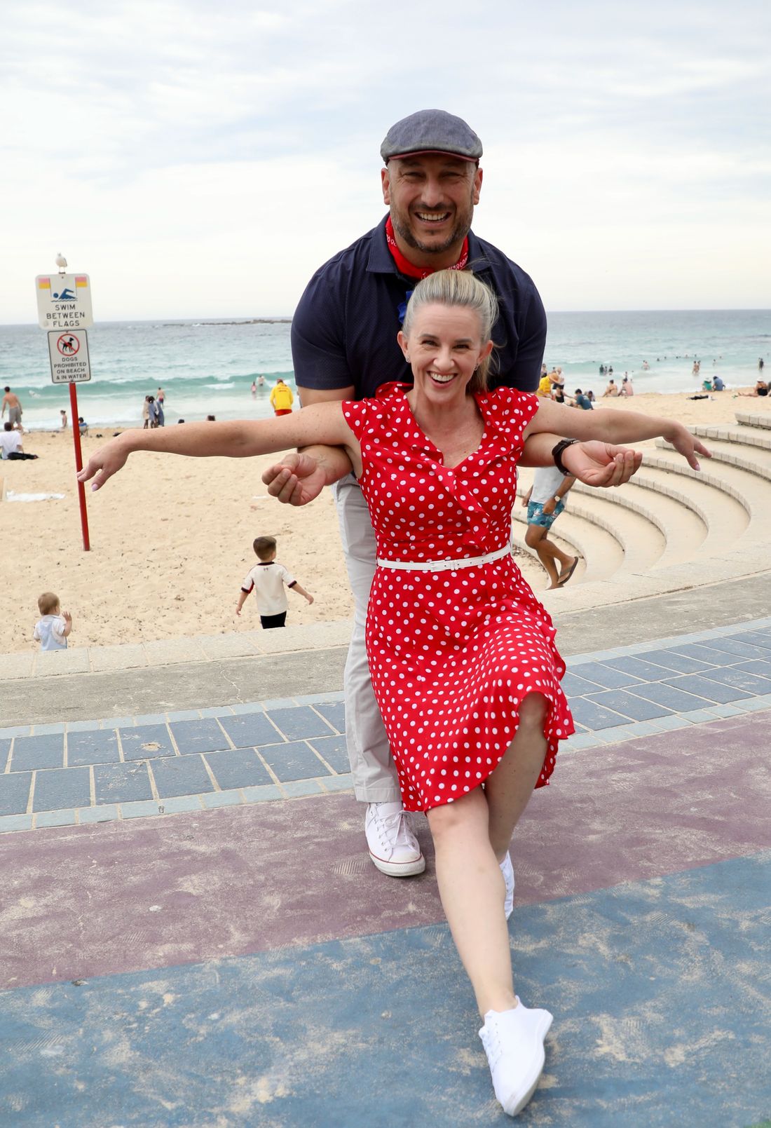 Mel and Marc on a beachfront promenade, arms flung wide mid-step, both laughing.