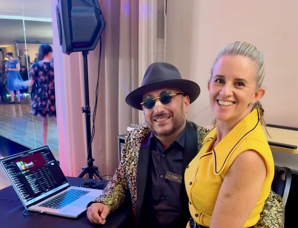Marc in a fedora and round sunglasses at the DJ table with a laptop, Mel beside him in a yellow dress, dancers reflected in the studio mirror behind them.