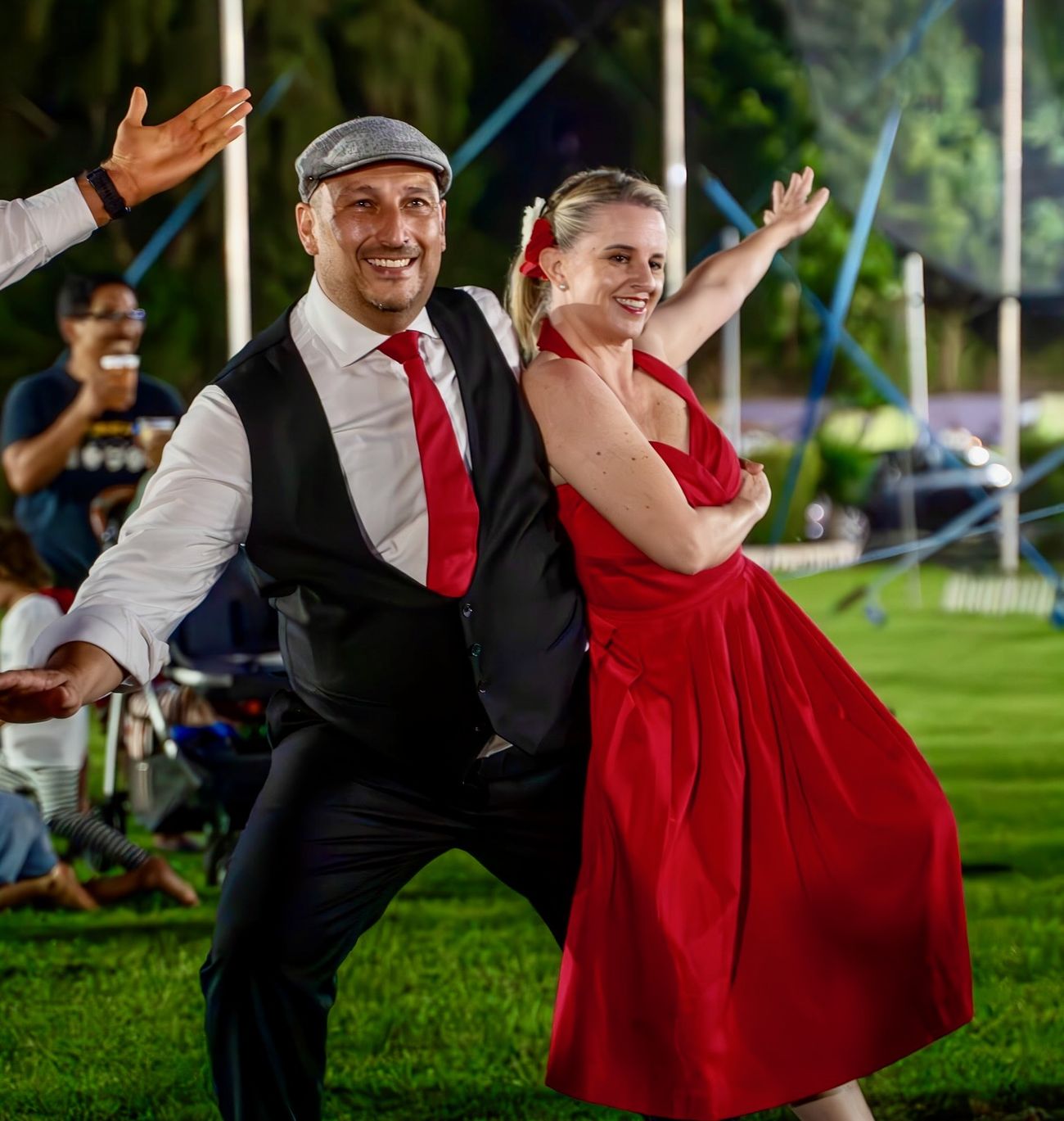 Mel and Marc mid-dance on grass at an outdoor event, both grinning with arms flung wide.